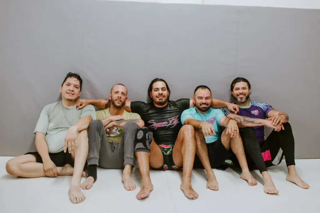 Five smiling adult students sitting against the mat wall after a No-Gi training session at SuperFly BJJ in Decatur