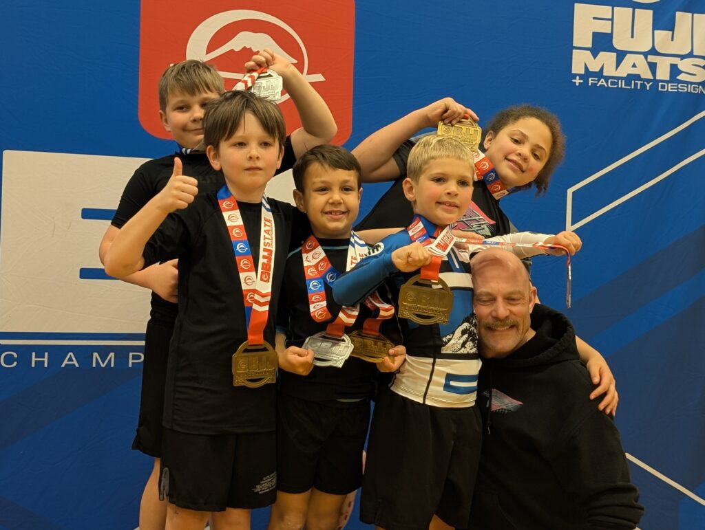 Youth martial arts students from Superfly BJJ in Decatur, GA, proudly displaying gold and silver medals with their coach after a Kids BJJ tournament.