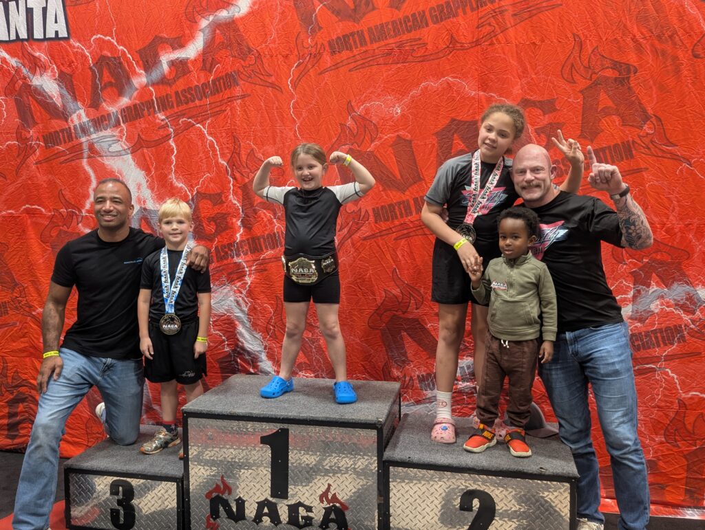 Kids Brazilian Jiu-Jitsu medal winners celebrating on NAGA tournament podium with coaches