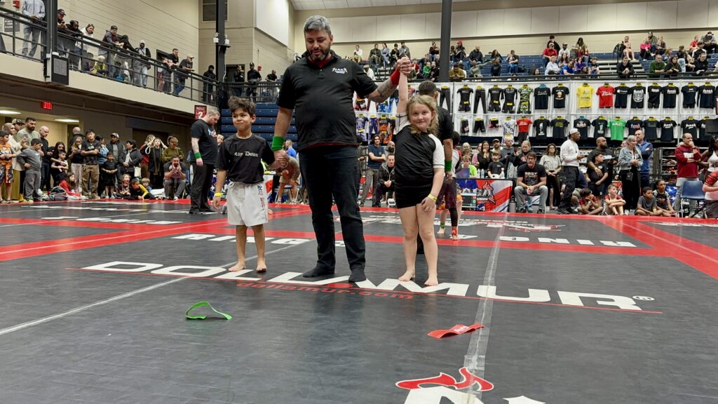 Kids BJJ tournament scene with referee awarding victory to a SuperFly BJJ competition team member by raising her hand after a match.